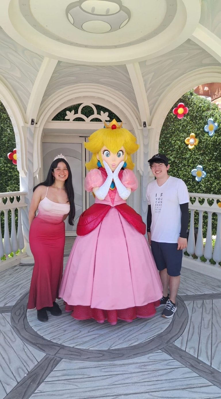 Two visitors pose with Princess Peach character in front of white pavilion with ivy walls and colorful flowers