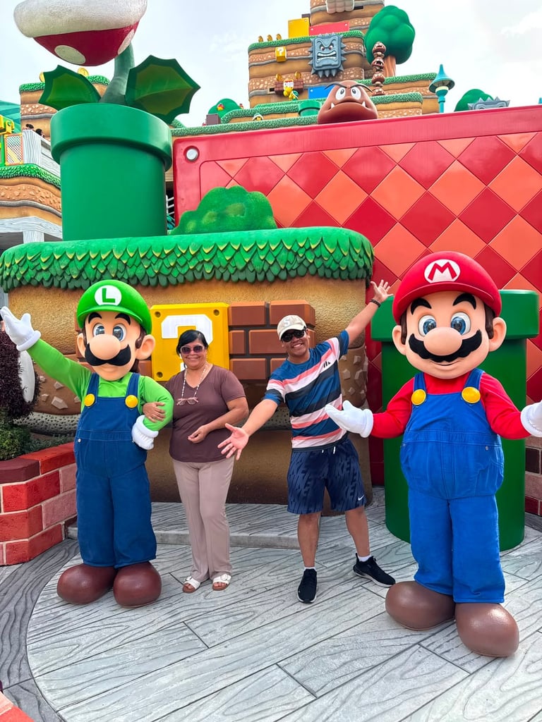 Two visitors pose with Mario and Luigi character mascots at a colorful theme park attraction
